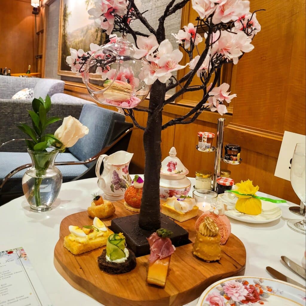 A wooden tray with assorted savory and sweet courses, centered around a cherry blossom tree centerpiece at Afternoon Spring Tea, Ritz Carlton Atlanta.