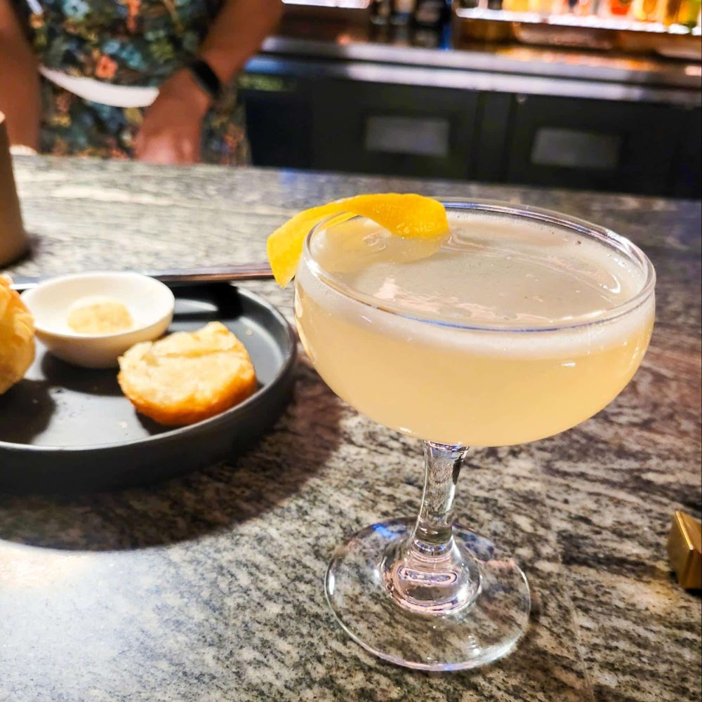 A cocktail garnished with a lemon twist served at the bar of Lazy Betty, accompanied by a small plate of appetizers.